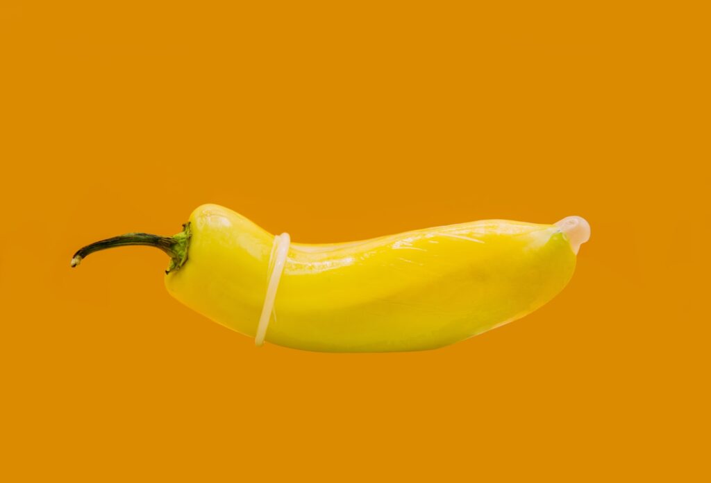 A single yellow banana pepper wearing a condom on a mustard yellow background.
