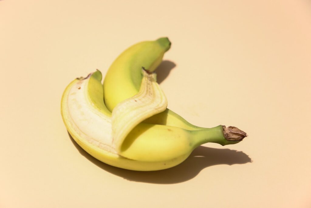 Pair of bananas, one partially peeled, on light yellow background.