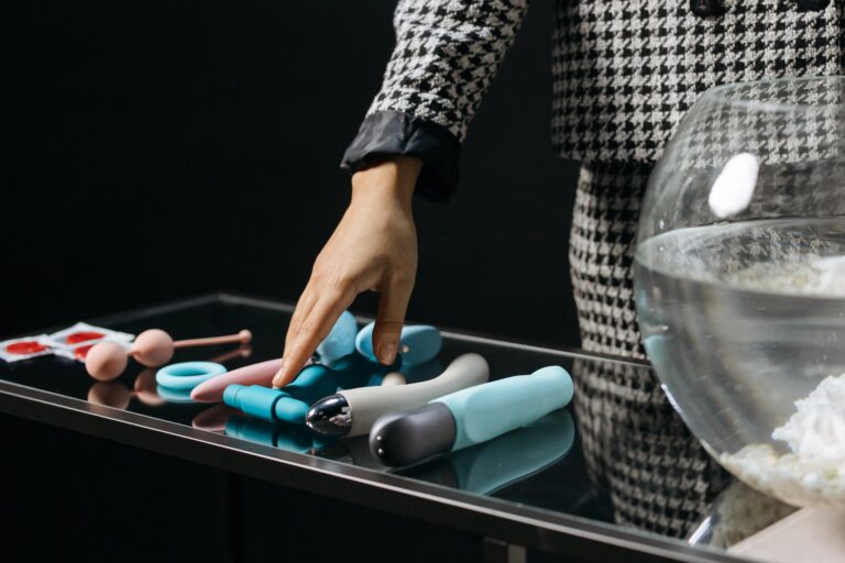 A variety of sex toys laid out on a glass table, as a hand moves over them.