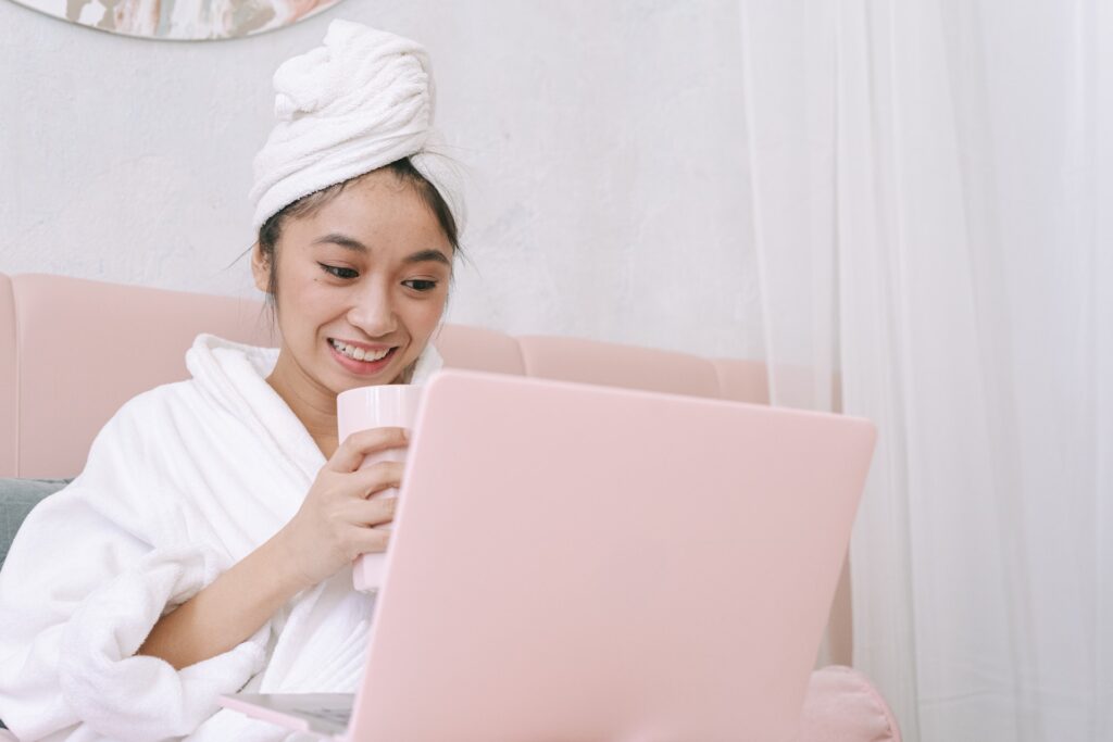 Woman smiles coyly at blush pink computer, with hair wrapped in towel, drinking coffee.