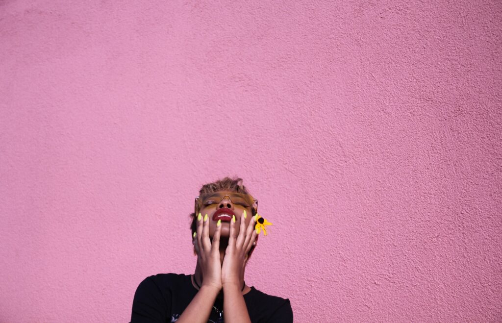 Woman with bright yellow fingernails touches her uplifted face against pink background.