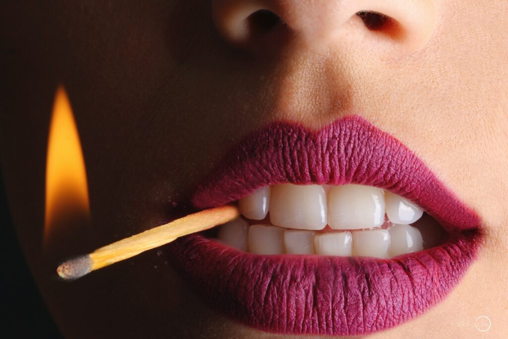 Woman holding lit match between her teeth, wearing plum matte lipstick