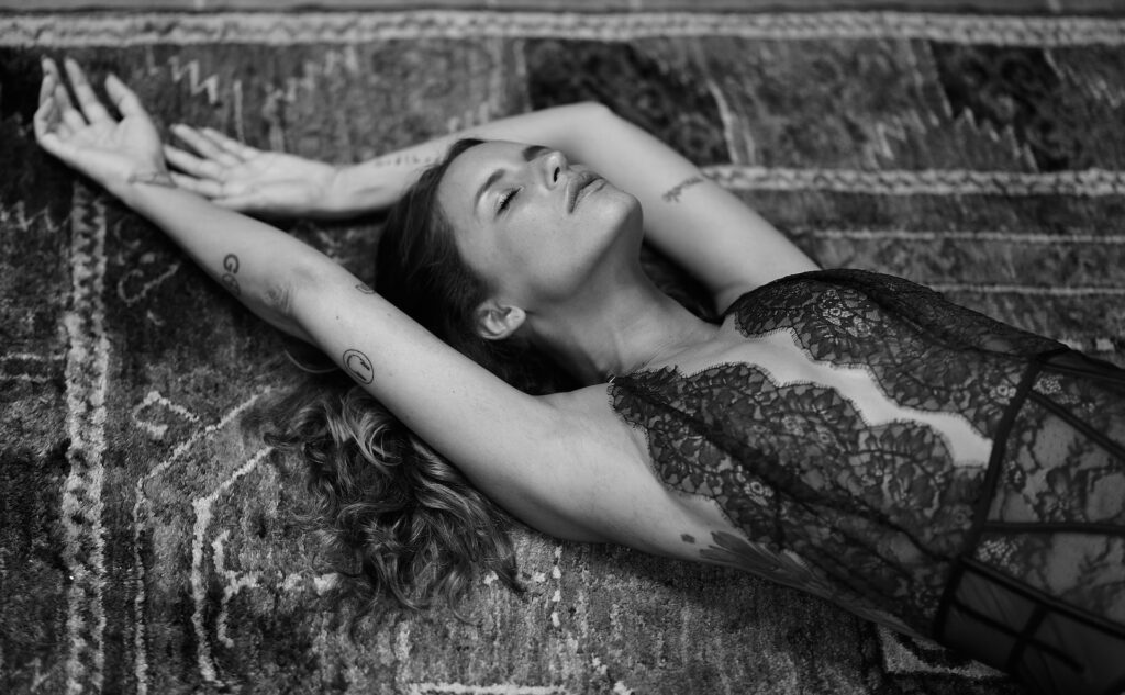 Woman drapes herself ecstatically across a kilim rug in low-cut lace camisole, eyes closed.