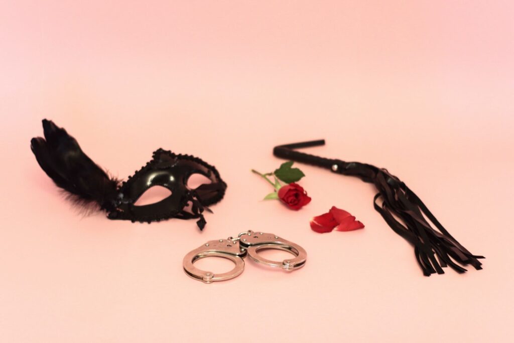 Black mask, black flogger, and metal handcuffs with single rose on light pink background.