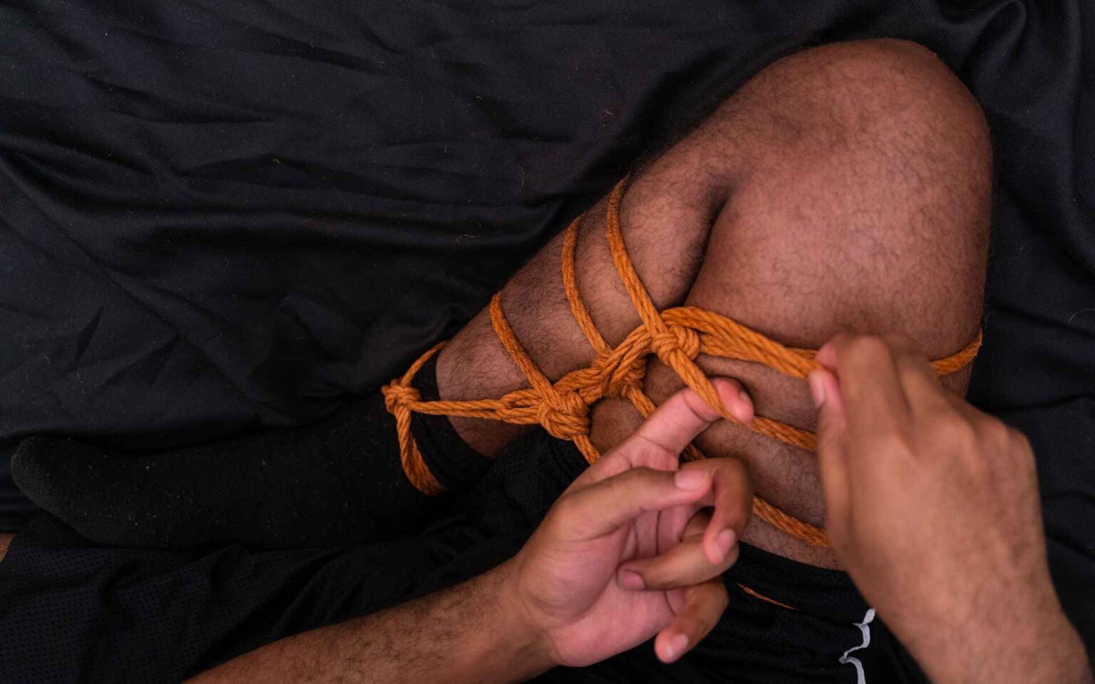 Man’s lower and upper leg tied together with orange Shibari rope on black background