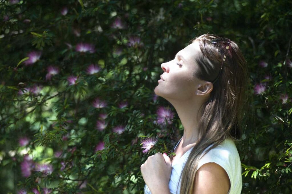 Woman serenely closes eyes as the sun hits her face, standing against a bush with purple flowers