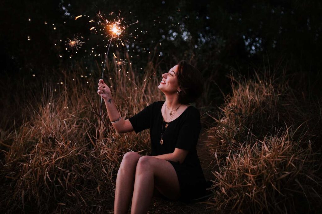 Woman sitting outdoors with lit sparkler, grinning with joy