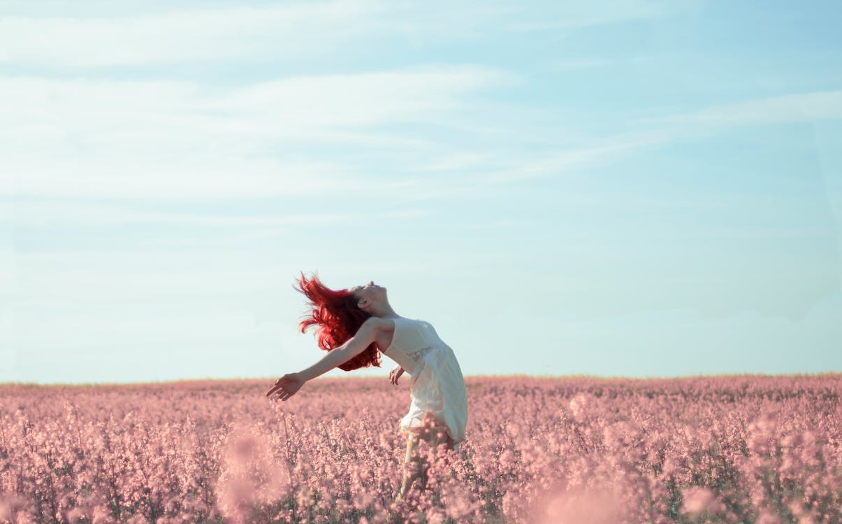 Woman with bright red hair throws arms open in ecstasy in field of pink flowers against blue sky.