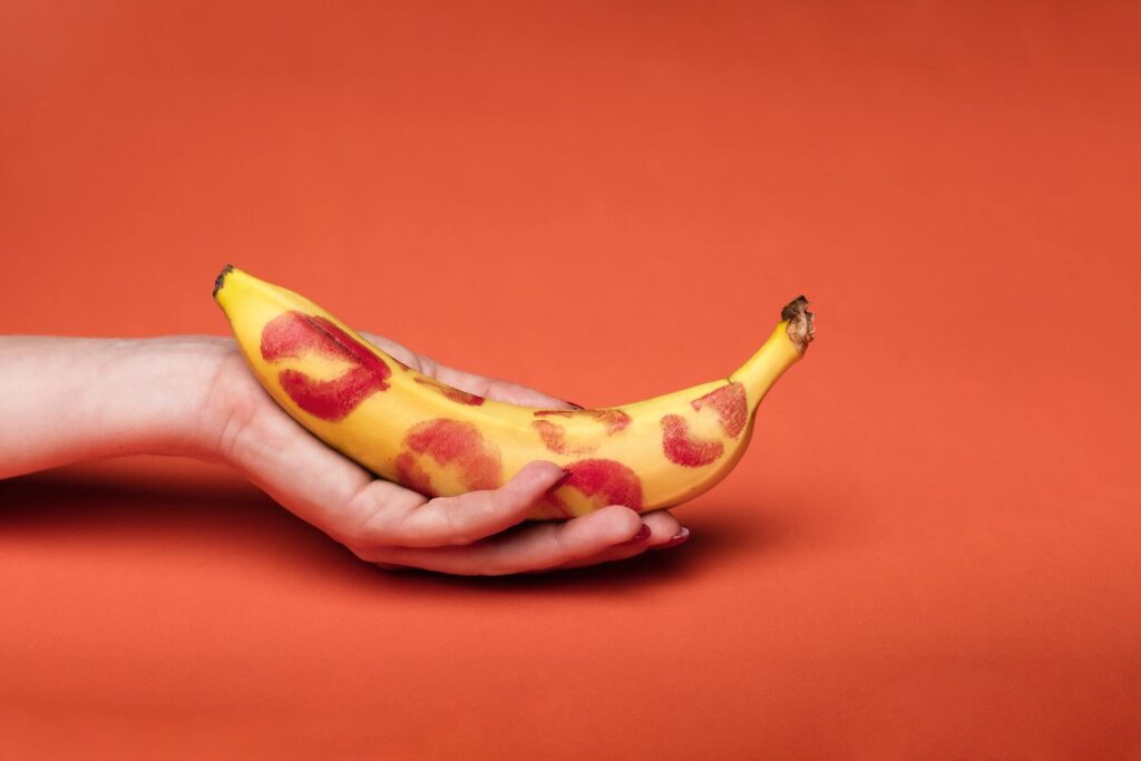 Hand holds banana covered in red lipstick kisses against red background.