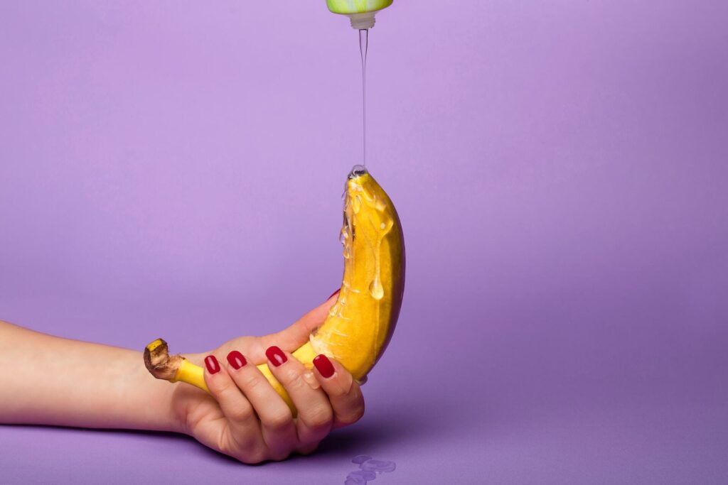 Hand with painted, red nails holds banana while lube is poured over it.
