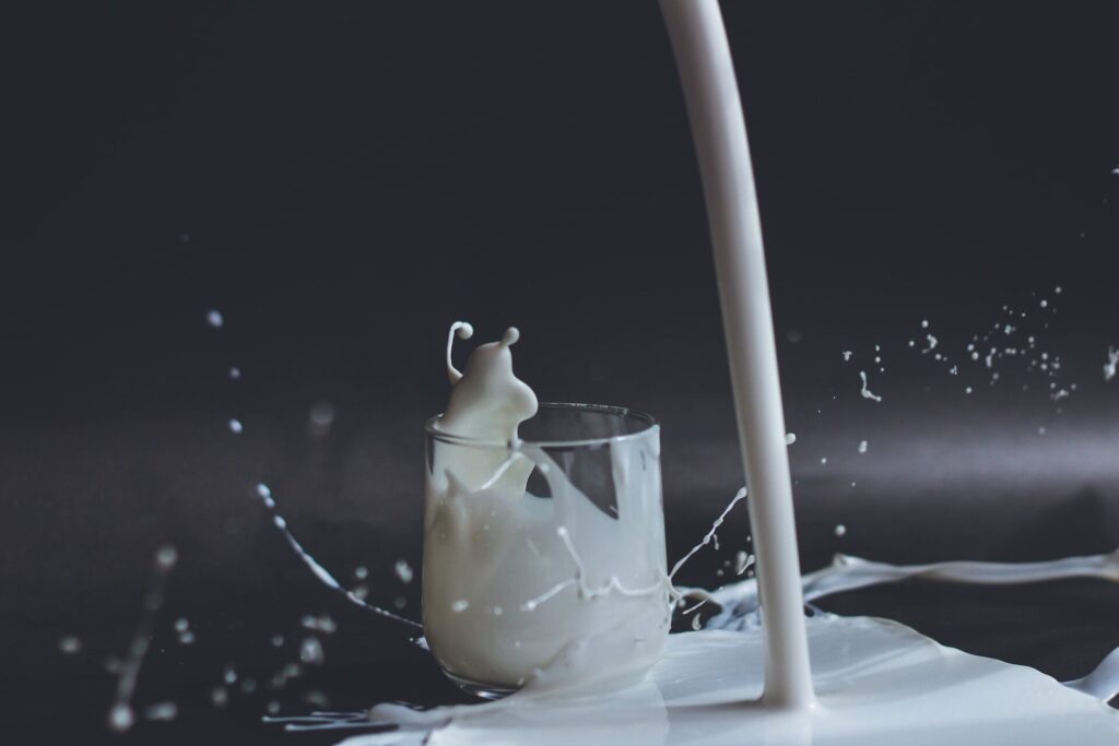 Stream of milk spills over black table next to glass filled with splashing milk.
