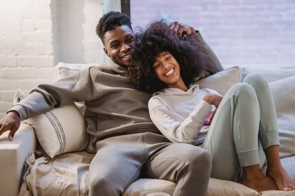 Smiling, content couple cuddling on neutral-tone couch against white brick wall.