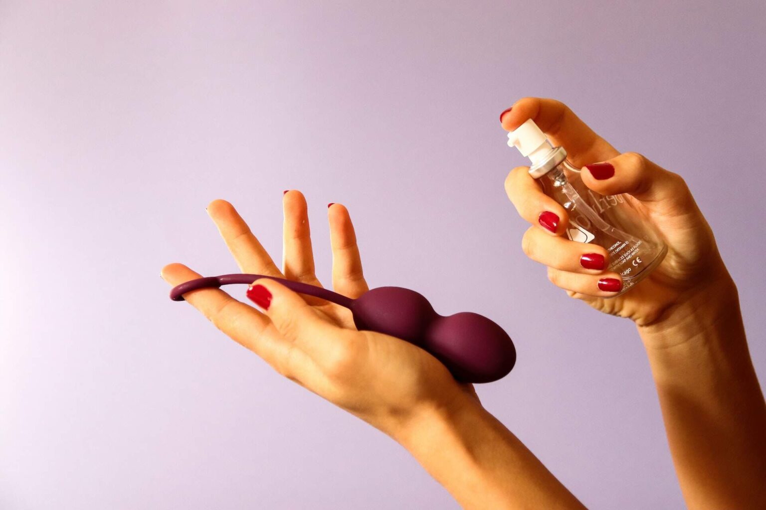 A hand holds a sex toy, while the other hand prepares to spray it with sex toy cleaner against a lavender background.