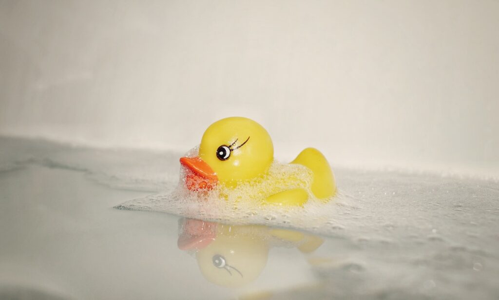 Soapy rubber duck floats on top of bath water in white bathtub.