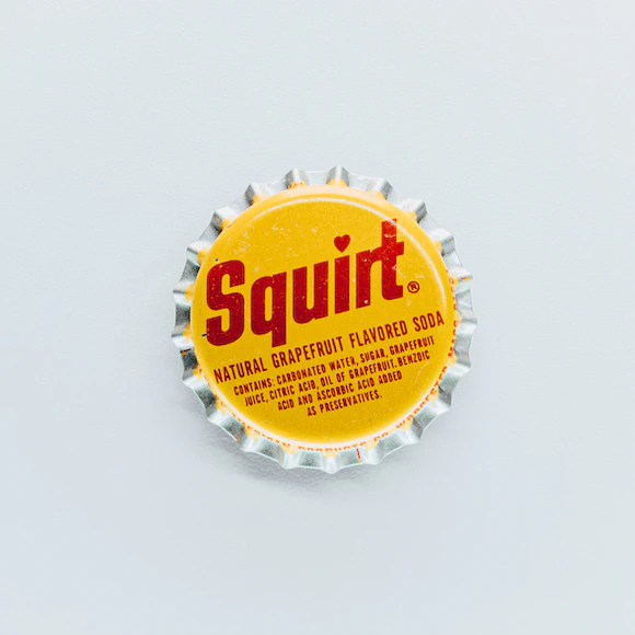 Bottle cap from “Squirt” soda, in yellow with red lettering on gray background.