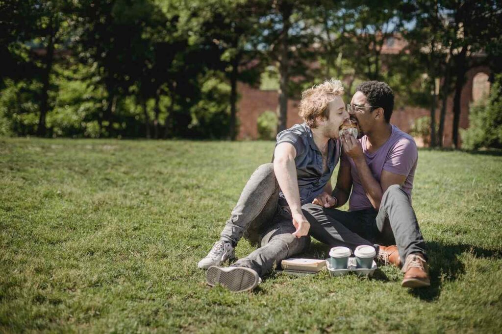 Two men sitting on the grass, enjoying a flirtatious snack together.