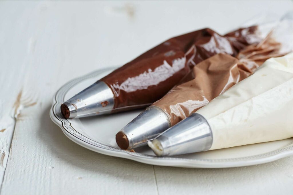 Three piping tubes with metal tips filled with dark, milk, and white chocolate icing.