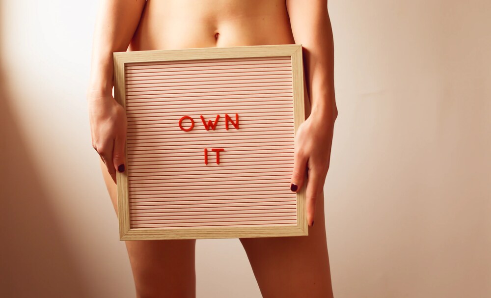 Nude woman holds sign reading, “Own it”.